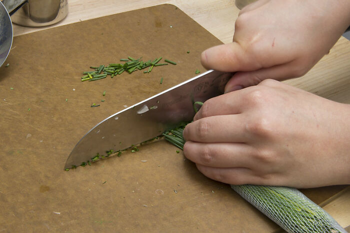 Hands chopping fresh herbs on a wooden cutting board with a large kitchen knife, showcasing a cooking tool product worth money.
