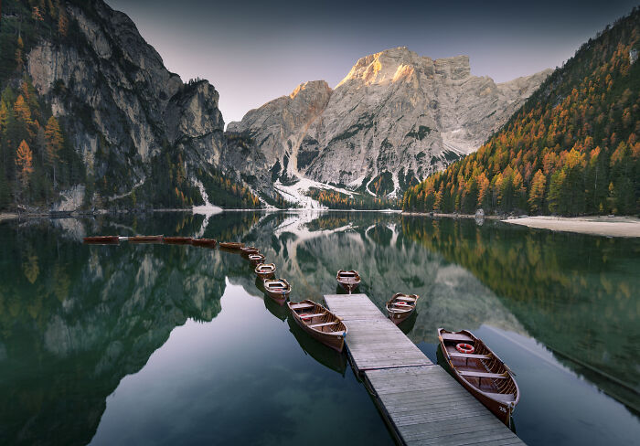 Wooden boats lined up at a dock on a calm lake surrounded by colorful fall trees and the Alps mountains at sunrise.