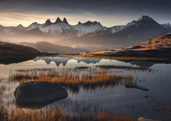 Mountain photography of the Alps during colorful fall with vibrant orange grasses reflecting in a calm lake at sunrise.