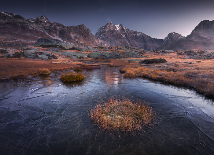 Colorful fall landscape in the Alps featuring frozen water, autumn grass, and rugged mountain peaks at dusk.
