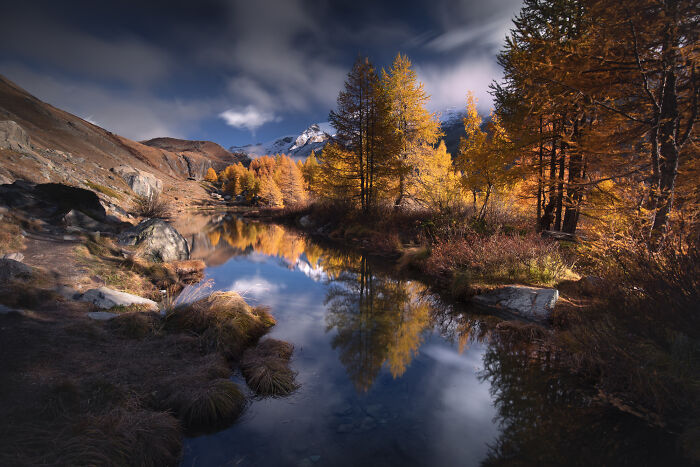 Colorful fall scene in the Alps with golden trees reflected in a calm river, captured by a mountain photographer.