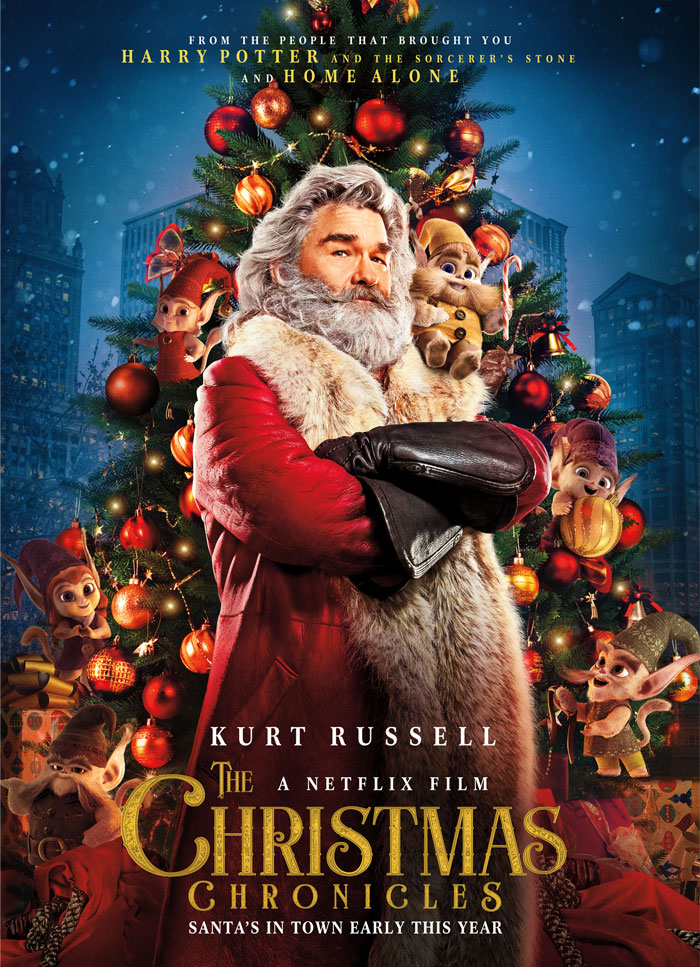 Kurt Russell as Santa in "The Christmas Chronicles" with elves, surrounded by Christmas decorations.