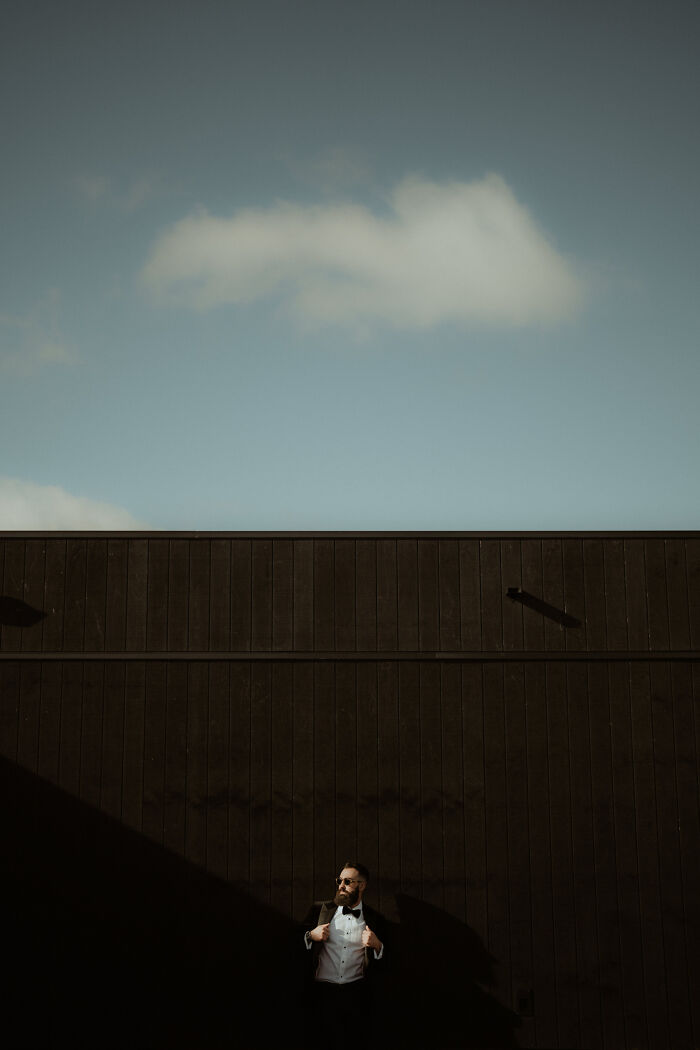Groom in a tuxedo posing against a dark wall under blue sky, featured in international wedding photographer submissions.