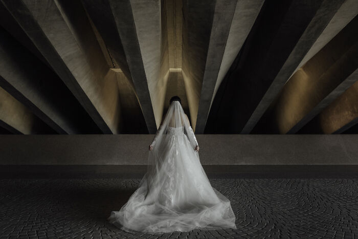 Bride in a flowing white wedding dress and veil standing under dramatic architectural beams, showcasing wedding photography.