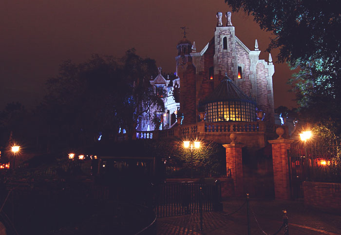 Dark nighttime view of a Disney theme park castle with illuminated windows and surrounding trees and lamps.