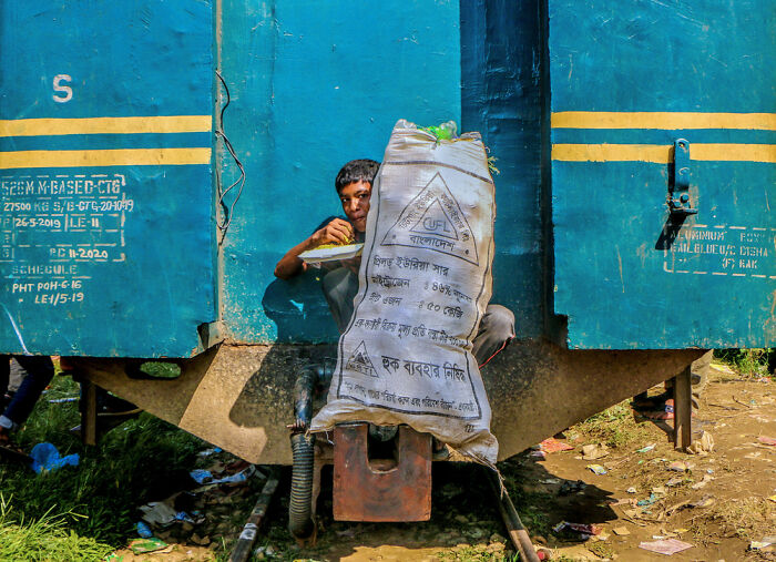 Man eating while sitting between blue train cars, highlighting environmental photography and human impact themes.