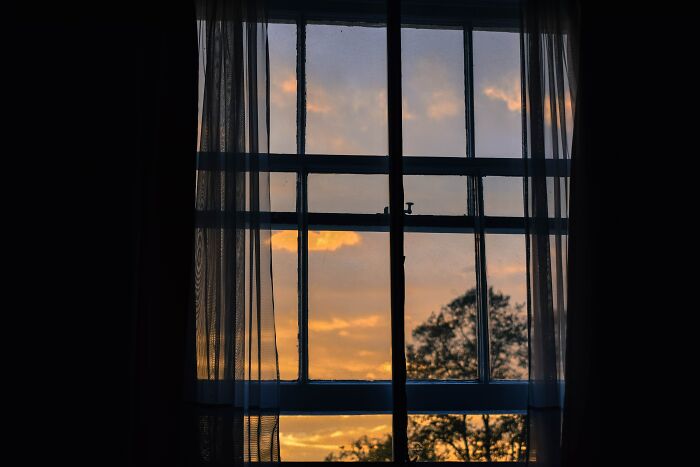 View of a window at sunset with sheer curtains, illustrating advice on where you should never hide your stuff from burglars.