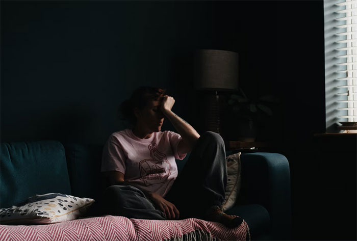 Person sitting alone in a dark room by a window, reflecting on life after being labeled gifted as a child.