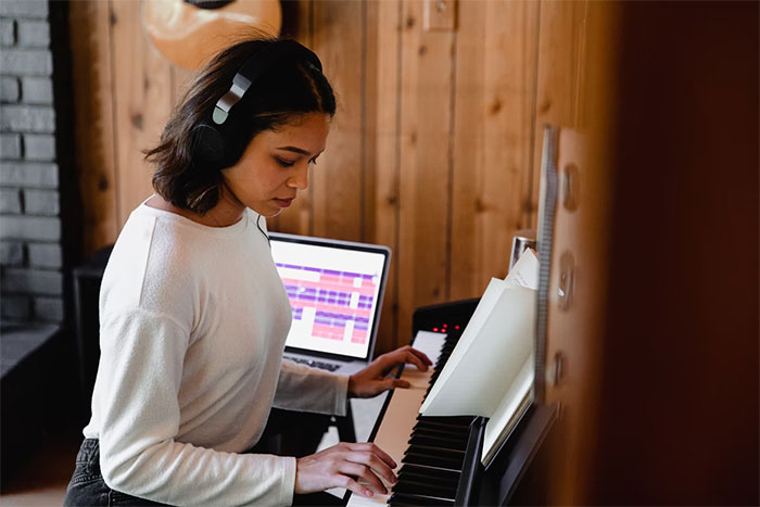 Young gifted woman practicing piano with headphones on in a cozy room, focused on music and laptop nearby.