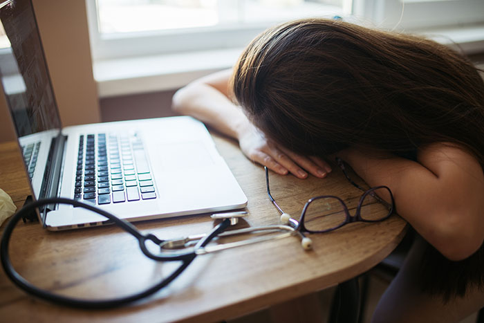 Person labeled gifted with head down on table next to laptop, stethoscope, and glasses showing fatigue or stress at work.