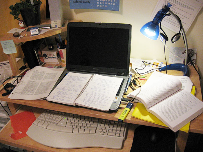 Cluttered study desk with laptop, notebooks, textbooks, and a blue desk lamp illustrating gifted student workspace.