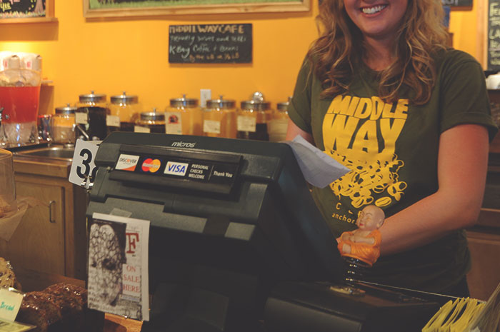 Smiling woman working behind a cafe counter with a cash register, representing good deeds kept in secret.