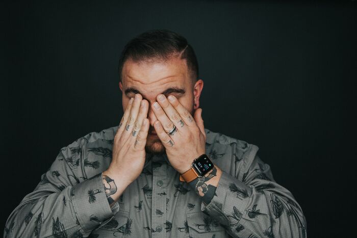 Man with tattoos and rings, wearing a patterned shirt and smartwatch, covering his face in a dark setting, social tips concept.