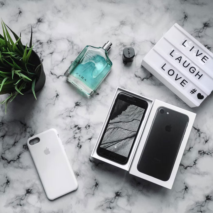 Flat lay of modern home trends items including smartphone, plant, fragrance, and lightbox on marble surface.