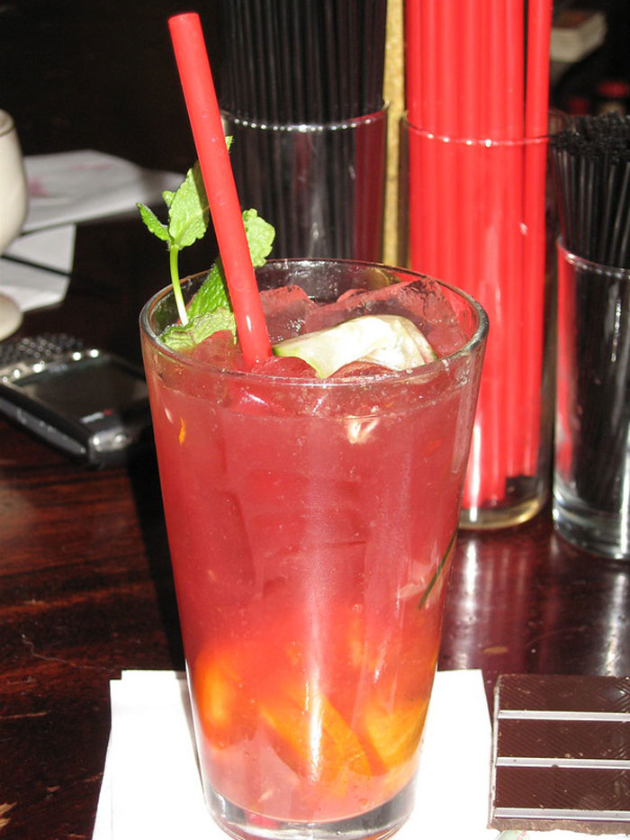 Red cocktail with ice, mint garnish, and a red straw on a bar counter, highlighting bartenders discussing drink orders.