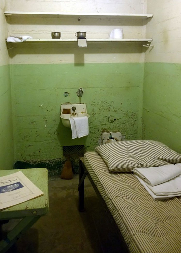 A small prison cell with a striped bed, a sink, shelves, and basic prison items representing incarceration life.