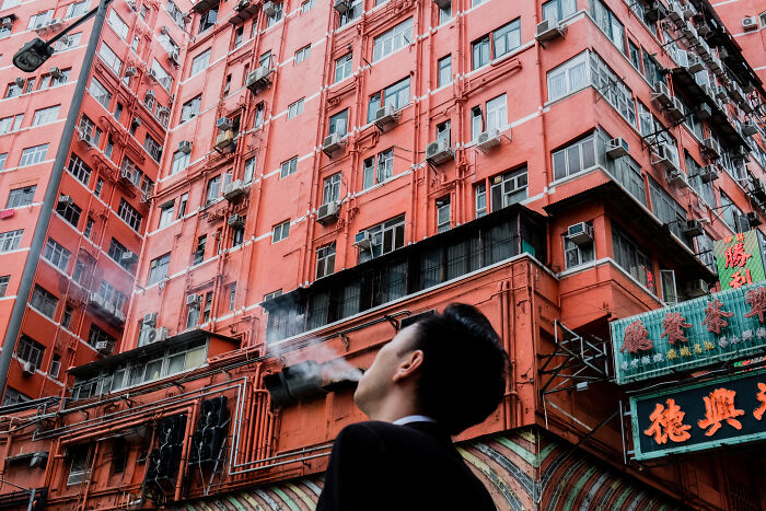 Man exhaling smoke in front of a vibrant red building, captured in a powerful street photo with urban details.