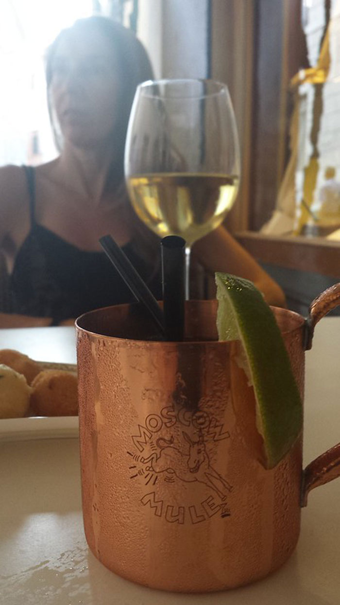 Copper Moscow Mule mug with lime wedge and black straws on table, blurred person and wine glass in background, bartenders drink orders.