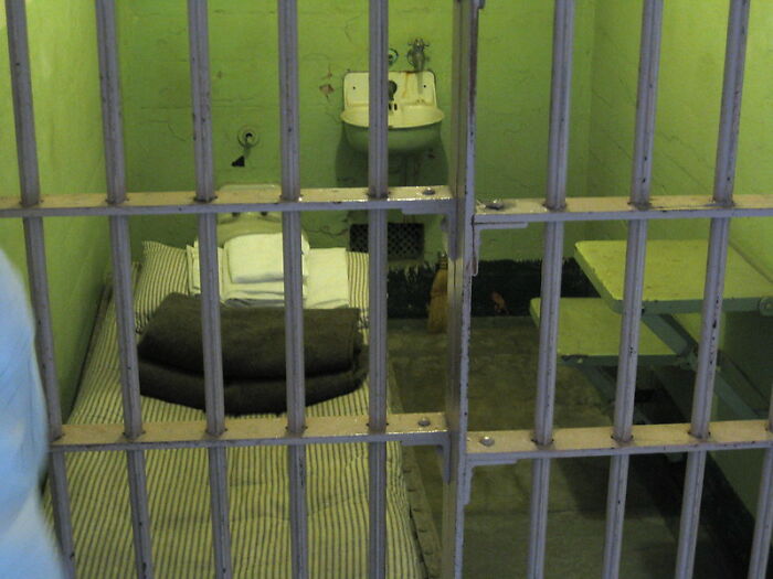 Prison cell interior with bed, folded blanket, sink, and small table behind metal bars representing incarceration experience.