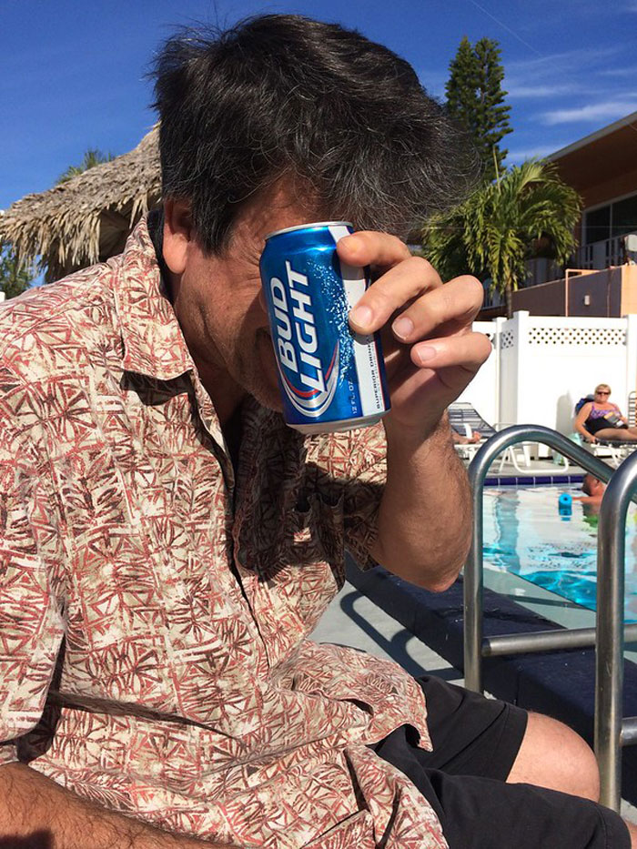 Man in patterned shirt by pool holding Bud Light can in front of face, illustrating bartenders online group drink order insights.