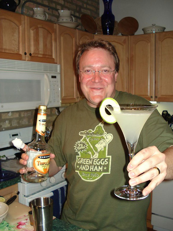 Man in kitchen holding cocktail glass with garnish and a bottle of Triple Sec liqueur, related to bartenders and drink orders.