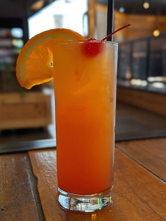 A colorful cocktail on a wooden table with an orange slice and cherry garnish, related to bartenders drink orders.