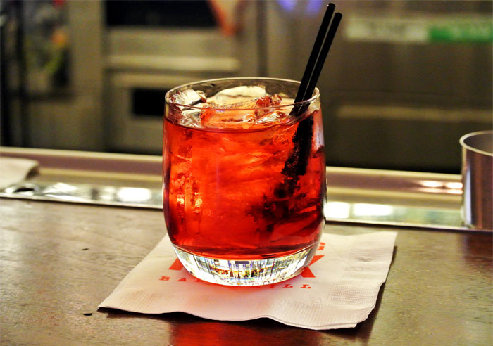 A vibrant cocktail drink with ice in a glass on a bar counter, highlighting bartenders discussing drink orders.
