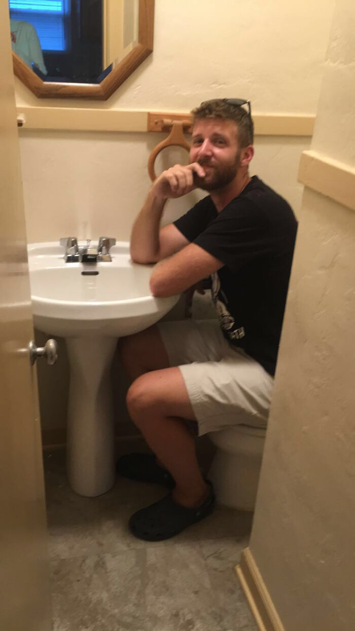 Person using a small bathroom with awkward interior design, sitting on a toilet placed closely next to a sink.