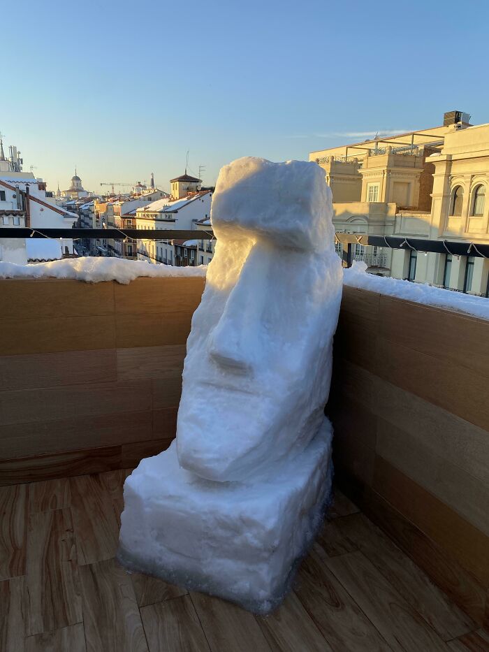 Snowman design resembling an Easter Island statue carved from snow on a rooftop with city buildings in the background.