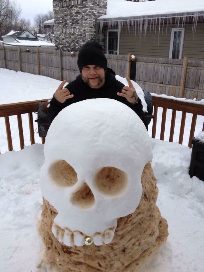 Man posing behind a creative snowman design shaped like a large skull with detailed features on a snowy deck.