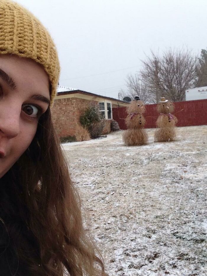 Woman in yellow knit hat taking selfie with unique straw snowman designs in snowy yard outdoors.