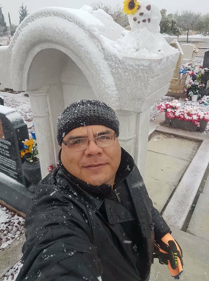 Man wearing hat and glasses takes selfie near creative snowman design with sunflower headpiece in snowy cemetery setting.