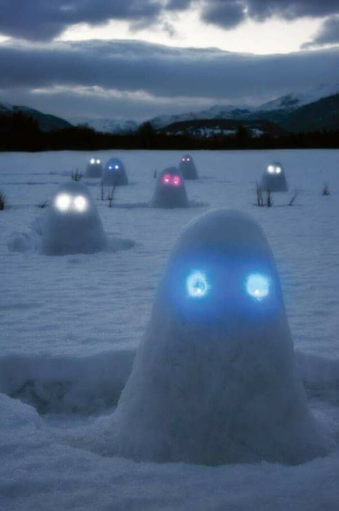 Glowing eyes on unique snowman designs in a snowy landscape at dusk with mountains in the background.