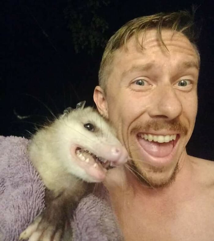 Person smiling with a possum wrapped in a towel, both making funny faces at the camera.