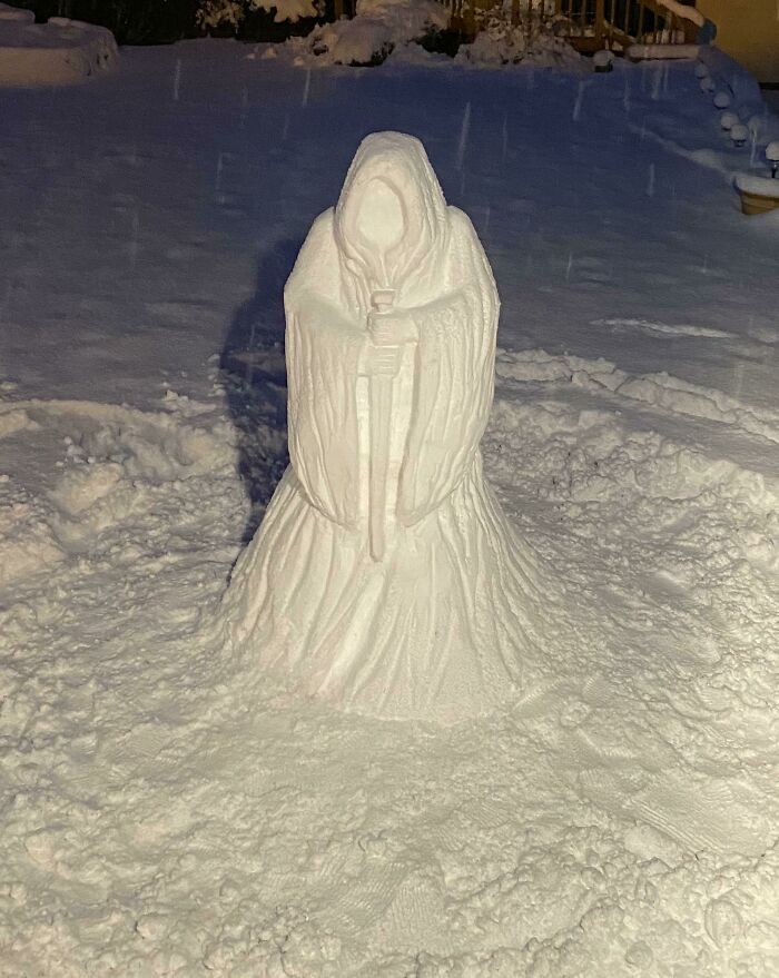 Snowman design sculpted to look like a cloaked figure holding a staff, standing in a snowy yard at night.
