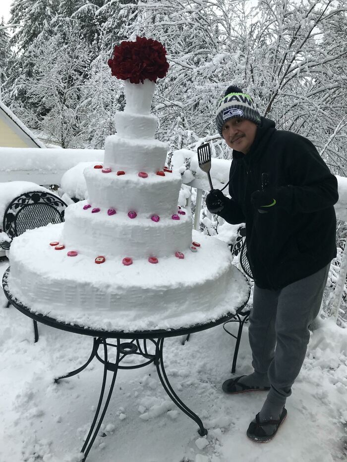 Man posing next to a creative multi-tiered snowman with red decorations in a snowy outdoor setting.