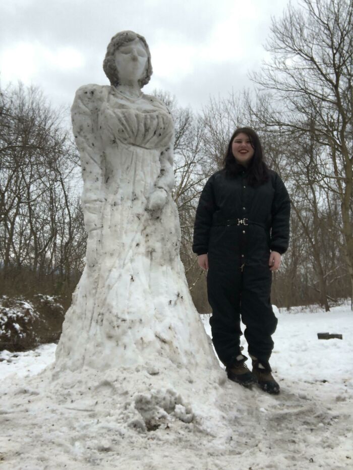 Tall snowman design sculpted as an elegant figure with detailed features standing next to a smiling person outdoors in winter.