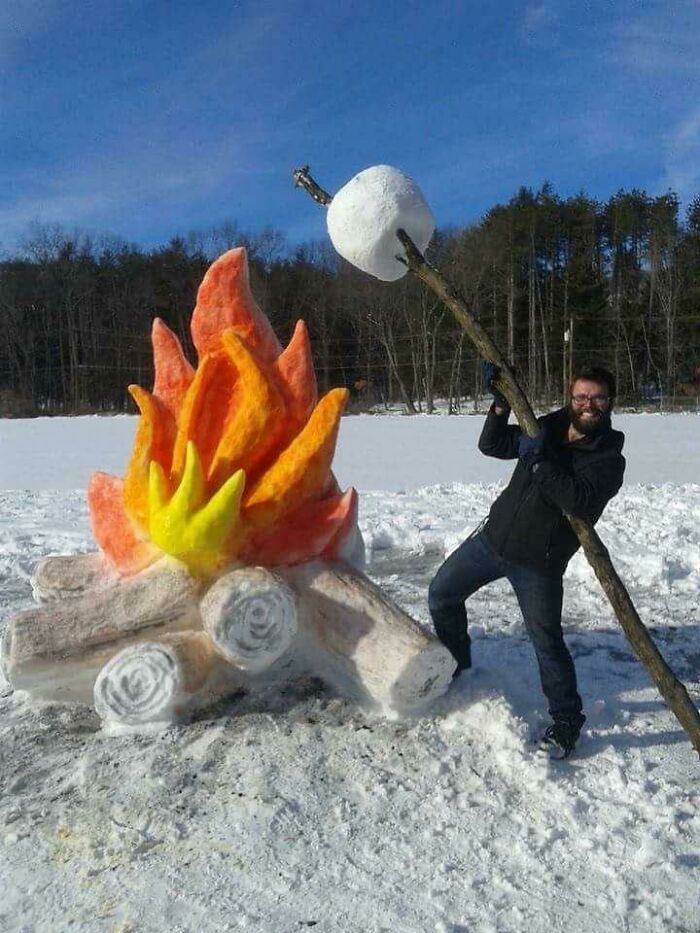 Man holding a stick with a large marshmallow near a colorful snow sculpture of a campfire in snowman designs.