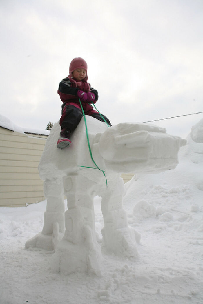 Child in winter clothing riding an elaborate snowman design shaped like a four-legged animal in a snowy yard.