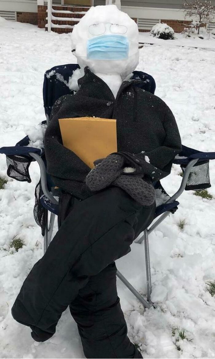 Snowman design sitting in a chair wearing a mask, glasses, dark clothes, and holding a large envelope in snowy yard.