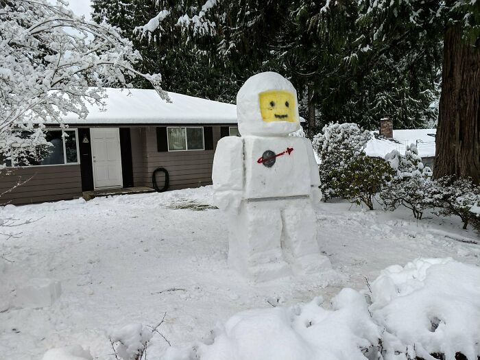 Snowman design shaped like a Lego astronaut figure made from snow in a snowy yard with trees and a house.