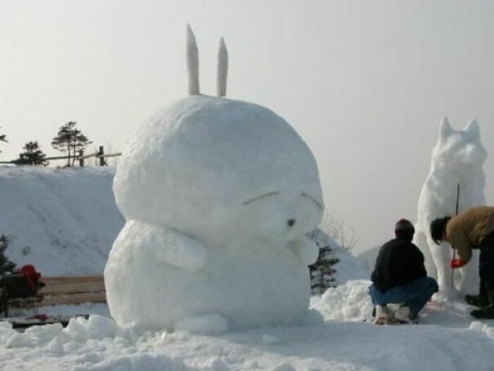 Large unique snowman design resembling a cartoon character with two people sculpting nearby in a snowy setting.