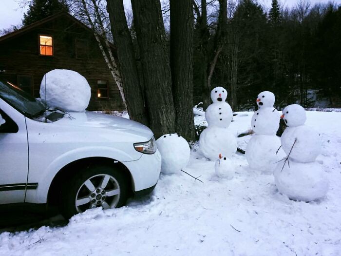 Creative snowman designs made from snowballs arranged around a car and trees in a snowy outdoor setting.