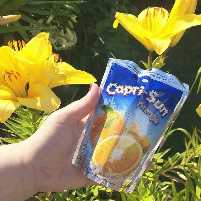 Hand holding Capri Sun orange drink, a nostalgic 90s food, against bright yellow flowers.