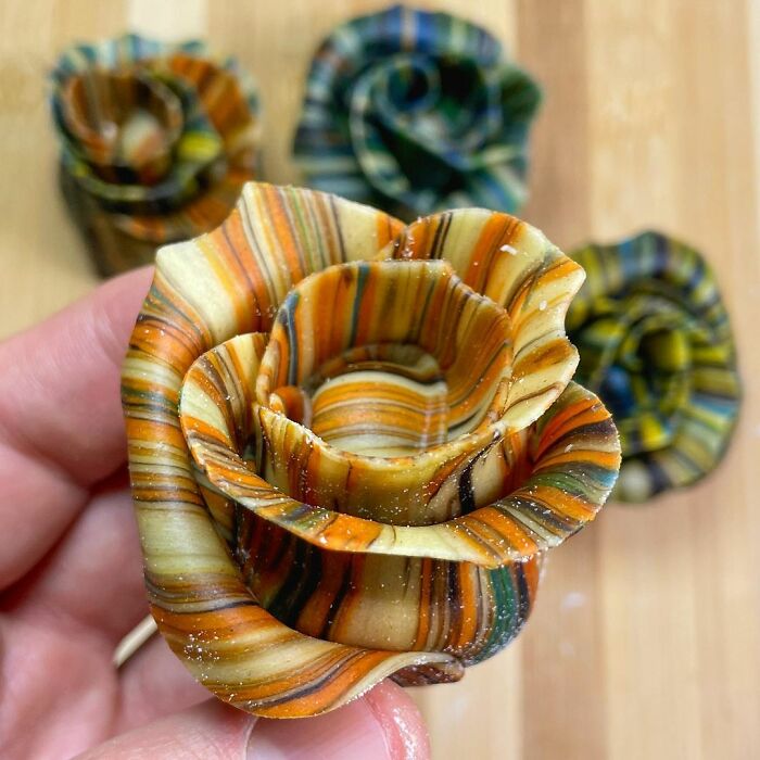 Hand holding a unique multicolored pasta design shaped like a rose with several similar pasta roses blurred in the background.