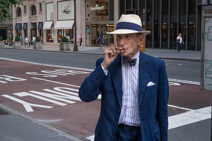 Elderly man in a blue suit and hat smoking a cigar on a city street in a powerful street photo setting.