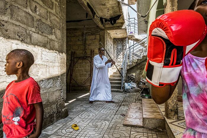 Young boy in red shirt, man playing flute in white robe, and person wearing red boxing gloves in urban street photo.