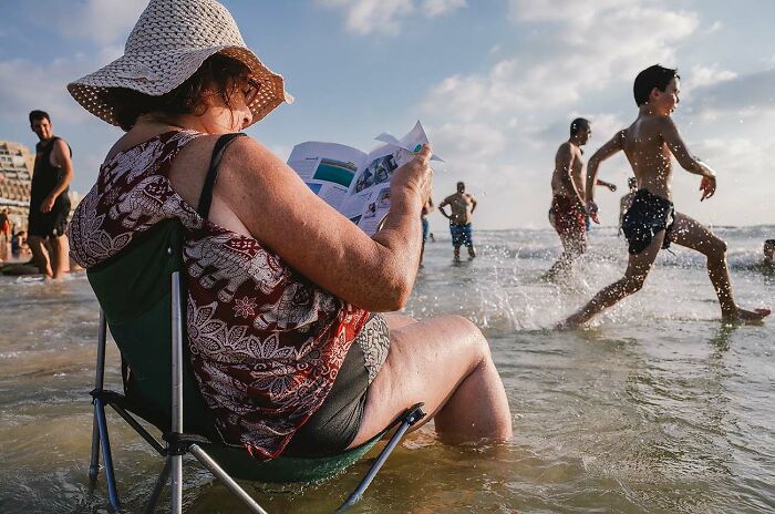 Woman wearing a sunhat sitting in a chair reading at the beach while children play in the water, powerful street photo.