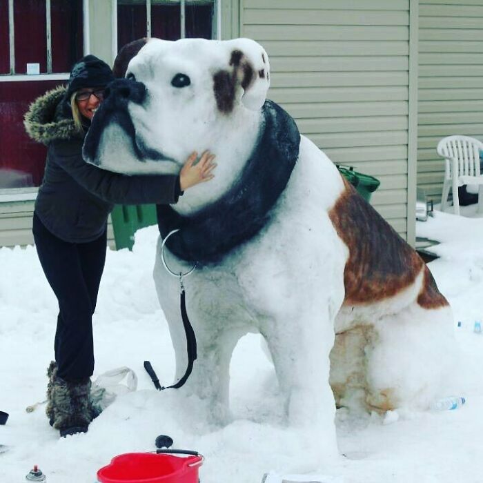 Woman posing with a large snowman design shaped like a realistic dog outside a house in winter.