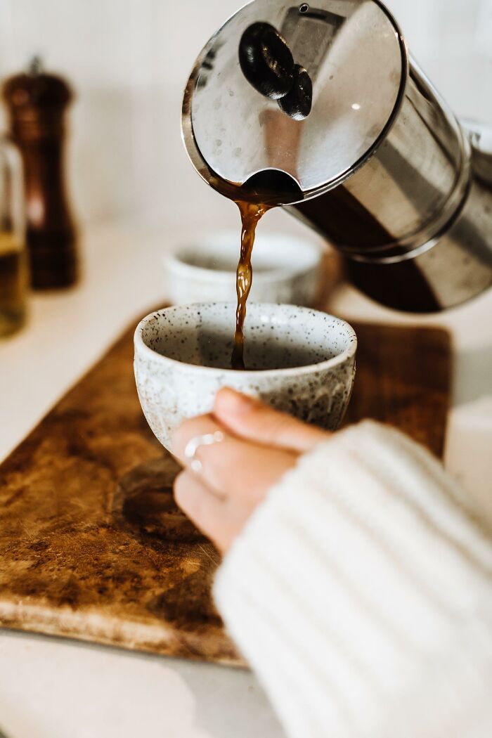 Coffee being poured from a stainless steel pot into a speckled cup, with a hand holding the cup on a wooden board.